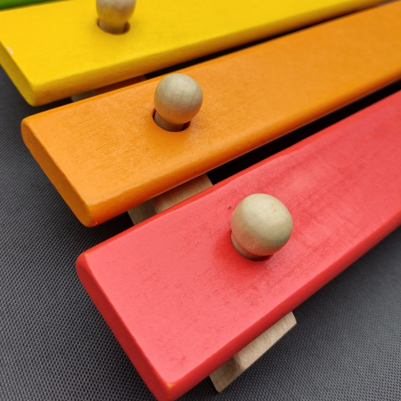 Op de afbeelding: Een houten xylofoon met gekleurde latten en ronde houten hamers. De latten zijn geverfd in tinten geel, oranje en rood.