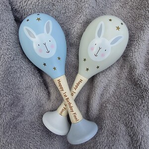 May include: Two baby maracas with bunny faces and gold stars. One is light blue, the other is light gray. The wooden handles read "Happy 1st Birthday Finn!" in a cursive font. The maracas are on a gray, fuzzy surface.