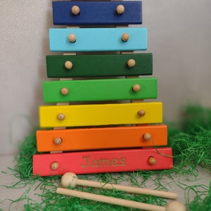 Op de afbeelding: Een regenboogkleurige houten xylofoon met een gepersonaliseerde naam, "James", gegraveerd op de onderste balk. De xylofoon heeft twee houten hamers.