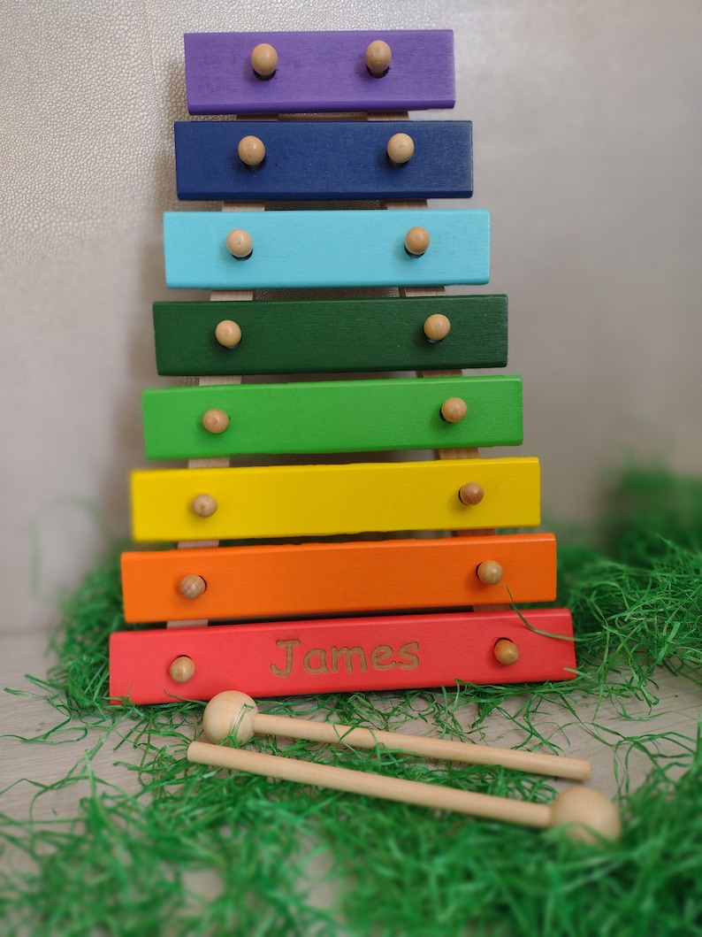 Op de afbeelding: Een regenboogkleurige houten xylofoon met een gepersonaliseerde naam, "James", gegraveerd op de onderste balk. De xylofoon heeft twee houten hamers.