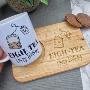 May include: A white ceramic mug and wooden coaster set. The mug has a tea bag graphic and the text "EIGH-TEA Happy Birthday." The coaster has a matching tea bag design and the same text. Two biscuits are on the coaster.