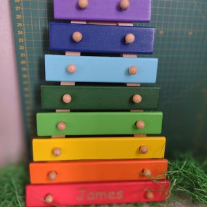Op de afbeelding: Een regenboogkleurig houten xylofoon met de gepersonaliseerde naam "James" gegraveerd op de onderkant. De xylofoon heeft houten hamers en is gemaakt van zeven verschillende gekleurde staven.