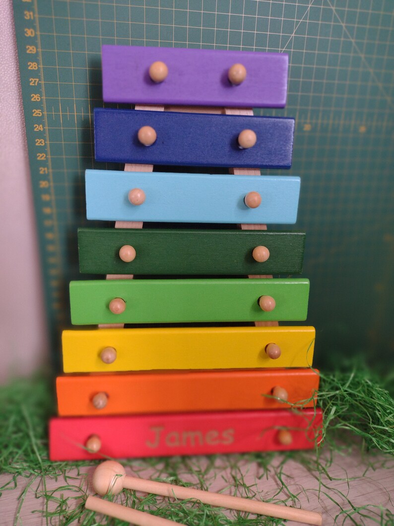 Op de afbeelding: Een regenboogkleurig houten xylofoon met de gepersonaliseerde naam "James" gegraveerd op de onderkant. De xylofoon heeft houten hamers en is gemaakt van zeven verschillende gekleurde staven.