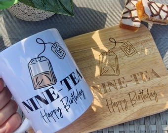 May include: White ceramic mug with the text "NINE-TEA Happy Birthday" and a tea bag graphic. A wooden cutting board with the same text and tea bag graphic is next to the mug. A pastry is on the cutting board.