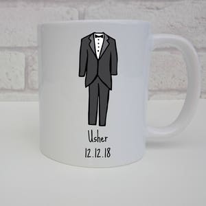 Può includere: Tazza in ceramica bianca con un'illustrazione in bianco e nero di una persona in smoking. Il testo "Usher 12.12.18" è stampato sotto l'illustrazione.
