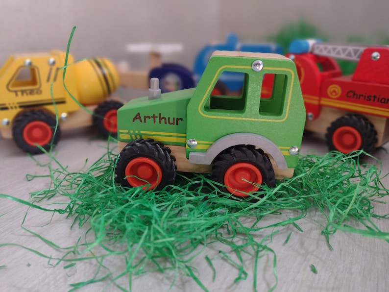 May include: A green wooden toy tractor with the name "Arthur" printed on the side. The tractor has black wheels with red centers and is sitting on green grass.