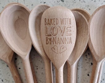 May include: A collection of wooden spoons, with one spoon engraved with the words "BAKED WITH LOVE BY NANNA" and two heart symbols. The spoons are made of light-colored wood and are arranged in a fan shape.