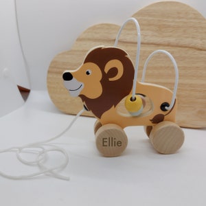 May include: A wooden pull-along lion toy with a white pull string. The lion has a tan body, brown mane, and a smiling face. It features a bead maze and the name "Ellie" printed on a wheel. The toy is designed for young children.