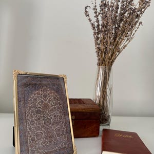 May include: A rectangular picture frame with a gold-toned metal border and ornate corner detailing, containing a print of a detailed floral tapestry pattern in shades of blue, gray, and brown, standing upright on a flat surface.
