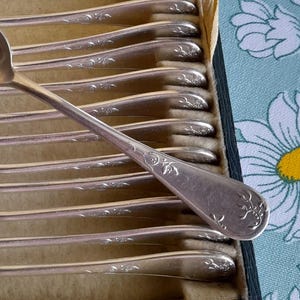 May include: A set of silver spoons in a vintage box. The spoons have a classic design with a floral pattern on the handles. The box is lined with a light green fabric and has a dark green exterior.