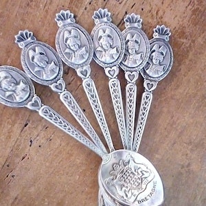 Pode incluir: Um conjunto de colheres prateadas com cabos detalhados e taças ovais. Cada colher apresenta um design decorativo de uma pessoa com chapéu. As taças das colheres exibem um brasão e a palavra "BRETAGNE".