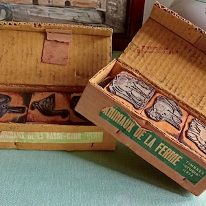 Könnte beinhalten: Zwei Vintage-Kartons mit Gummistempeln. Eine Box ist geöffnet und zeigt Stempel mit Tiermotiven. Die Boxen haben Etiketten mit französischem Text. Die Boxen befinden sich auf einer Tischdecke mit Blumenmuster.