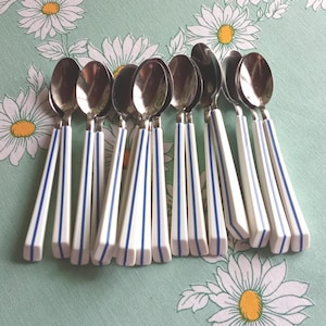 Puede incluir: Una colección de diez cucharas de acero inoxidable con mangos blancos con rayas verticales azules. Las cucharas están dispuestas sobre un mantel verde claro decorado con patrones de margaritas blancas.