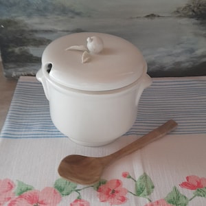 May include: White ceramic sugar bowl with a lid featuring a small apple design. The bowl is sitting on a white and blue striped kitchen towel with a floral pattern.