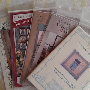 Peut inclure: Une pile de patrons de point de croix avec différents motifs, dont "Honeybees", "The Little", "Come What May" et "The Peaceful Home".