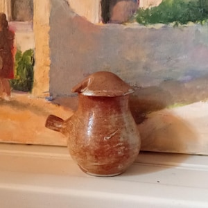 May include: A small, handmade ceramic pitcher with a brown glaze. The pitcher has a spout, a small handle, and a lid. It is sitting on a white surface in front of a painting of a European village scene.