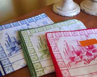 Puede incluir: Tres paños de cocina con fondo blanco y diseño azul, verde y rojo. Cada paño presenta una pequeña ilustración detallada de un castillo con techo rojo y un árbol verde. El paño rojo tiene una pequeña etiqueta naranja con el texto "TOUALLE DE CUISINE - MADE IN FRANCE - 50 x 70 CM".
