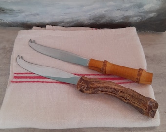 May include: Two cheese knives with wooden handles. One knife has a bamboo handle and the other has a brown, natural wood handle. The knives are on a white linen kitchen towel with red stripes.