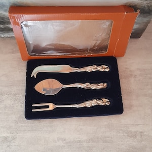 Pode incluir: Um conjunto de três facas de queijo prateadas em uma caixa. As facas têm um design floral decorativo e são embaladas em uma caixa forrada de veludo azul.