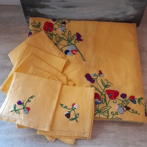 Könnte beinhalten: Ein Set gelber Leinen-Servietten mit gestickten Blumenmustern. Die Servietten sind gefaltet und auf einem Tisch gestapelt. Die Stickerei zeigt grüne Ranken, rote, violette und blaue Blumen.