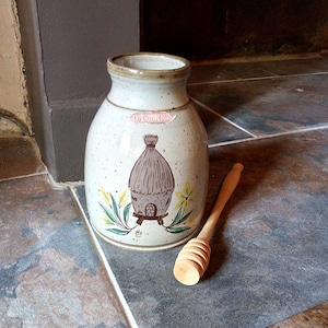 May include: A white ceramic honey pot with a brown beehive design and the text "Le Courtois" in pink. A wooden honey dipper is next to the pot.