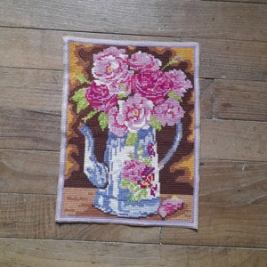 May include: A cross-stitch embroidery of a blue and white pitcher filled with pink and white flowers. The pitcher has a floral design and is sitting on a brown background.