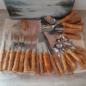 Pode incluir: Um conjunto de talheres com cabos de bambu. O conjunto inclui garfos, facas e colheres. Os talheres estão dispostos sobre um pano branco.