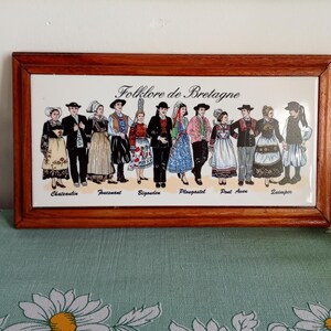 May include: A framed ceramic tile featuring a depiction of Breton folklore. The artwork showcases a line of figures in traditional costumes, with the text "Folklore de Bretagne" above. The frame is made of wood.