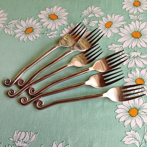 Peut inclure: Six fourchettes argentées vintage avec un design en spirale unique sur les manches. Les fourchettes sont disposées sur une nappe vert clair avec un motif de marguerites blanches. Les dents des fourchettes sont sombres.