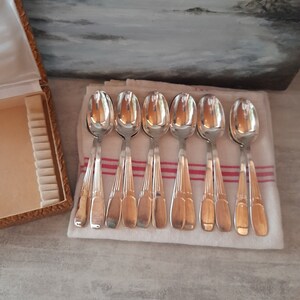 Pode incluir: Um conjunto de oito colheres de prata com cabos ornamentados, dispostas sobre uma toalha de linho branca com listras vermelhas. As colheres estão dispostas em duas filas de quatro, com os cabos voltados para o observador.