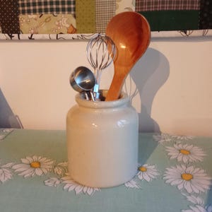 May include: A cream-colored ceramic kitchen utensil holder with a wooden spoon, a whisk, and a measuring spoon. The holder sits on a light blue tablecloth with a daisy pattern. A patchwork quilt hangs in the background.