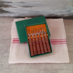 Peut inclure: Un ensemble de huit fourchettes en argent avec des manches en bois dans une boîte verte et orange. Les fourchettes sont disposées en rangée, les manches vers le haut. La boîte est posée sur une serviette blanche.