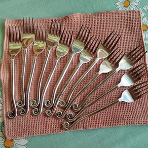 Puede incluir: Un conjunto de tenedores plateados con un diseño decorativo en espiral en la base del mango. Los tenedores están dispuestos sobre un paño texturizado de color marrón claro, sobre un fondo con estampado floral.