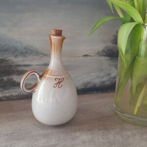 May include: A white ceramic oil bottle with a brown cork top and a brown handle. The bottle has a brown letter "H" on the front.