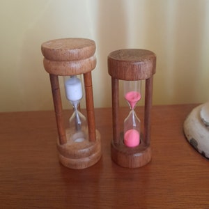 Può includere: Due clessidre di legno. La clessidra più grande ha sabbia bianca e quella più piccola ha sabbia rosa.