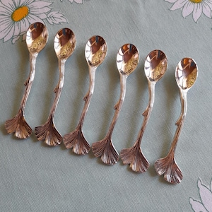 Könnte beinhalten: Sechs silberfarbene Löffel mit einem einzigartigen Design. Jeder Löffel hat eine kleine, ovale Schale und einen Stiel, der einem Ast ähnelt. Der Fuß jedes Löffels ist fächerförmig. Die Löffel liegen auf einer hellgrünen Oberfläche mit einem Gänseblümchenmuster.