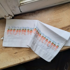 May include: White pillowcase with a delicate embroidered border of orange peppers and green leaves. The pillowcase is folded in half and lying on a wooden surface.