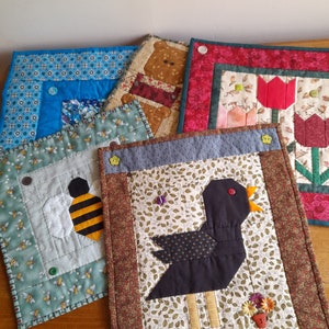 Può includere: Quattro arazzi da parete trapuntati fatti a mano con diversi disegni. Il primo è blu con un motivo geometrico, il secondo è verde con un'ape, il terzo è marrone con un orso e il quarto è rosso con un fiore.