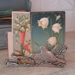 Puede incluir: Una figura de pájaro de metal plateado se encuentra frente a dos grabados de arte floral. Los grabados representan flores blancas en jarrones sobre un fondo azul y verde. La figura del pájaro es detallada y de estilo vintage.