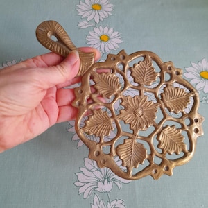 Puede incluir: Un salvamanteles de latón con un diseño decorativo de hojas y flores. El salvamanteles tiene un asa y es perfecto para proteger las superficies de los platos calientes.