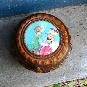 May include: A wooden music box with a round, ornate frame. The lid features a painted scene of two children, a boy and a girl, with the girl holding a bouquet of flowers. The box is decorated with a brown and gold finish.