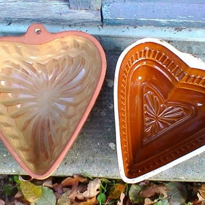 May include: Two heart-shaped ceramic baking molds. One is a light brown color with a raised design, the other is a darker brown with a white rim and a raised design.
