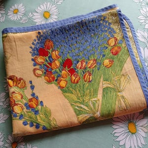 May include: Folded floral patterned fabric with a light yellow background. The fabric features bouquets of red and orange roses, blue flowers, and green stems tied with yellow ribbons. The edges are trimmed with a blue border.