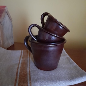 Puede incluir: Tres tazas de cerámica marrón apiladas una encima de la otra. Las tazas son de diferentes tamaños y tienen un aspecto rústico y hecho a mano.