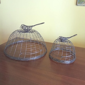Op de afbeelding: Twee draadsculpturen van vogelkooien, een grote en een kleine, elk bekroond met een draadvogel. De kooien zijn koepelvormig met een rasterpatroon en de vogels hebben gedetailleerde draadlichamen en lange staarten. De sculpturen staan op een houten oppervlak.