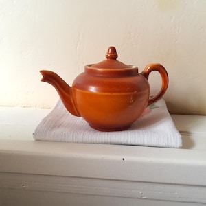 May include: A vintage, orange-brown ceramic teapot with a lid and handle, resting on a white cloth. The teapot has a rounded body and a curved spout. The background is a neutral, off-white color.