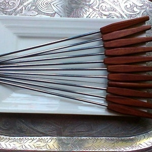 May include: A set of 10 stainless steel skewers with wooden handles. The skewers are arranged in a fan shape on a white plate. The handles are a rich brown color.