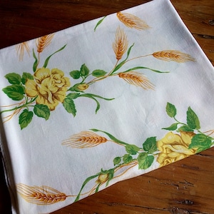 K&ouml;nnte beinhalten: Wei&szlig;e Tischdecke mit einem floralen Muster mit gelben Rosen und Weizen&auml;hren. Die Tischdecke hat einen wei&szlig;en Hintergrund mit gr&uuml;nen Bl&auml;ttern und gelben Weizen&auml;hren.