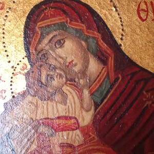 May include: A religious icon painting on a gold background featuring a figure in a dark red head covering holding a child. The child wears a light-colored garment with red accents. Greek lettering is visible in the upper corners of the panel.