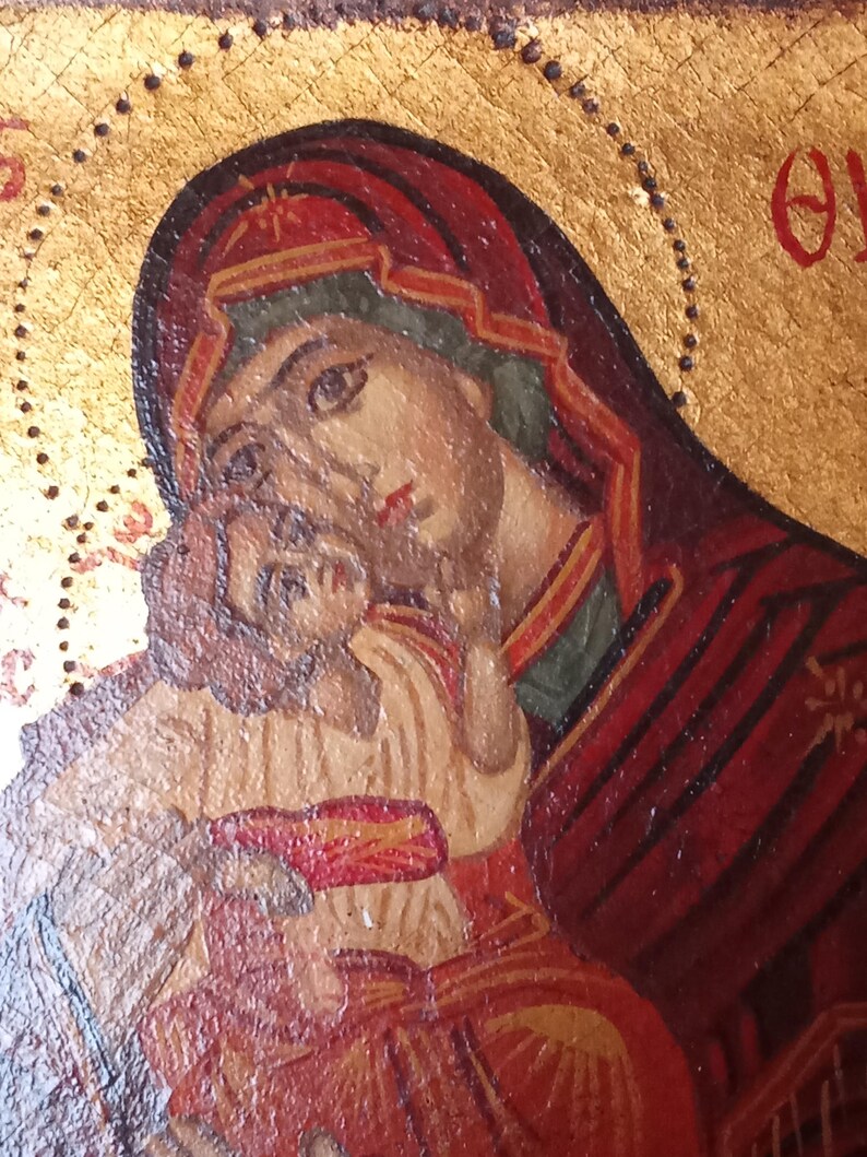 May include: A religious icon painting on a gold background featuring a figure in a dark red head covering holding a child. The child wears a light-colored garment with red accents. Greek lettering is visible in the upper corners of the panel.